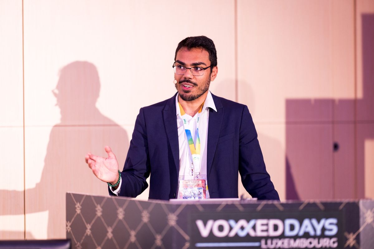 Sitraka Forler au pupitre de Voxxed Days Luxembourg 2025, badge d'intervenant au cou, la main ouverte pendant une explication